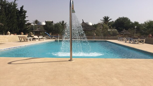 En swimmingpool med et springvand, omgivet af liggestole og palmer.