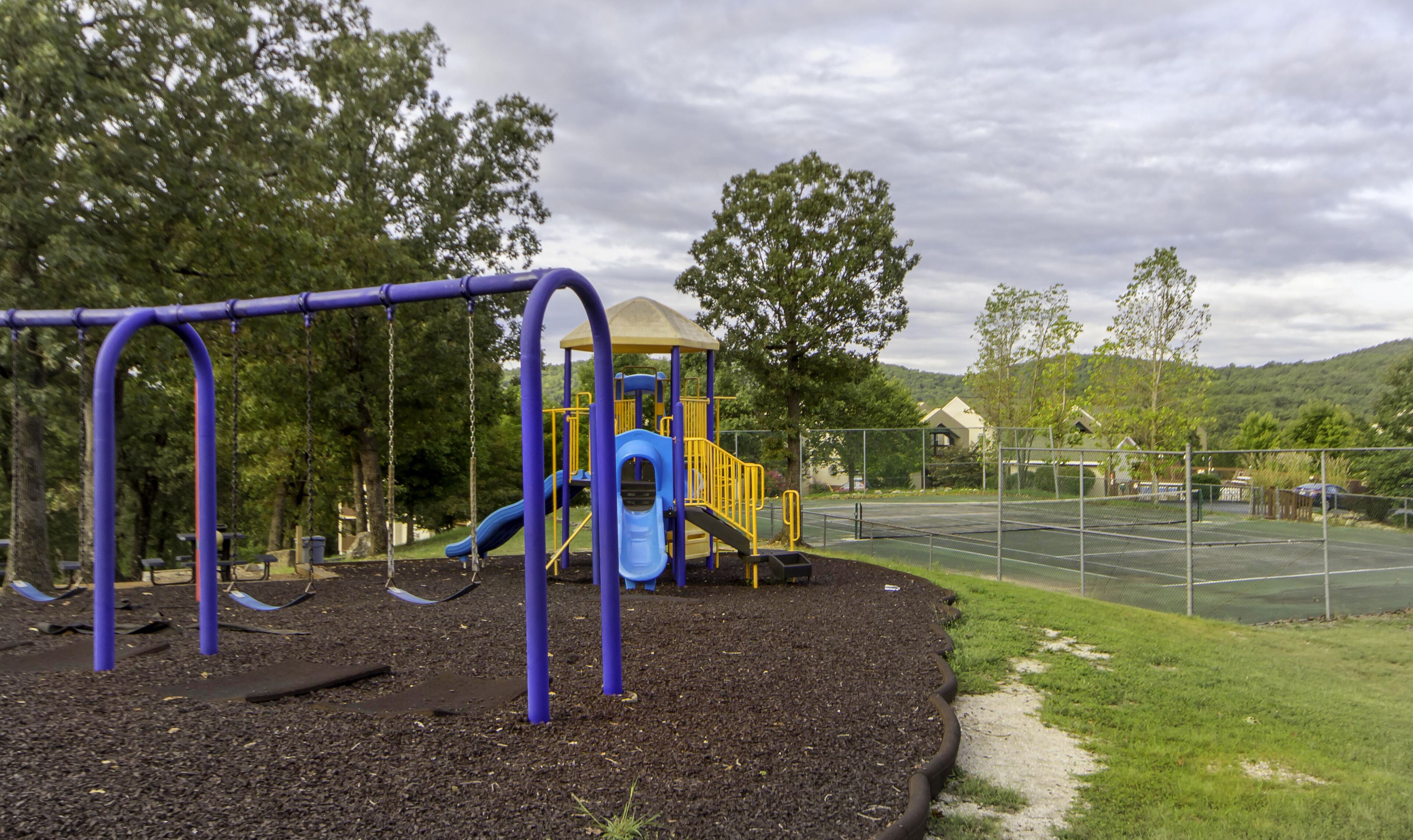 A playground with a slide and swings, surrounded by trees and a tennis court.