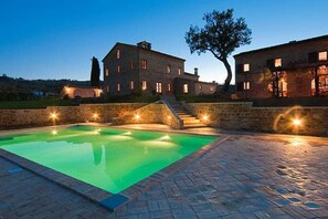 Un gran edificio de piedra con piscina, rodeado de un área pavimentada y un árbol.