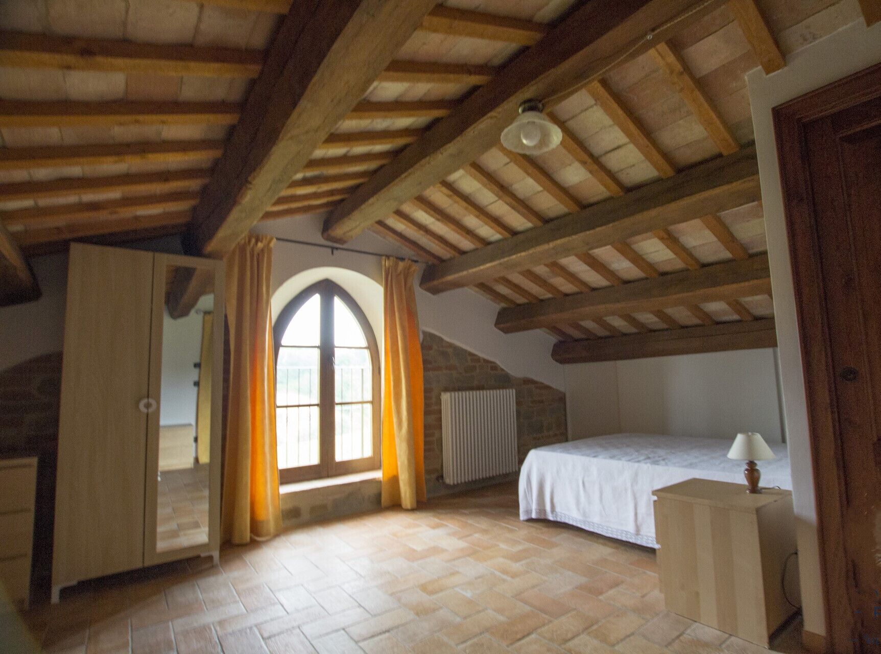 Une chambre avec un plafond en pente, une grande fenêtre en arc de cercle, un lit, une table de chevet et un radiateur.