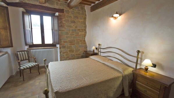 Un dormitorio con pared de piedra, una cama con cabecera, una silla, una mesita de noche con lámpara y una ventana con contraventanas.