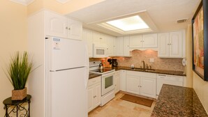 A kitchen with white cabinets, a refrigerator, a microwave, a stove, and a sink. There is a potted plant on a small table. - Condo with Gulf View just steps to our private Siesta Key Beach! (Sarasota)