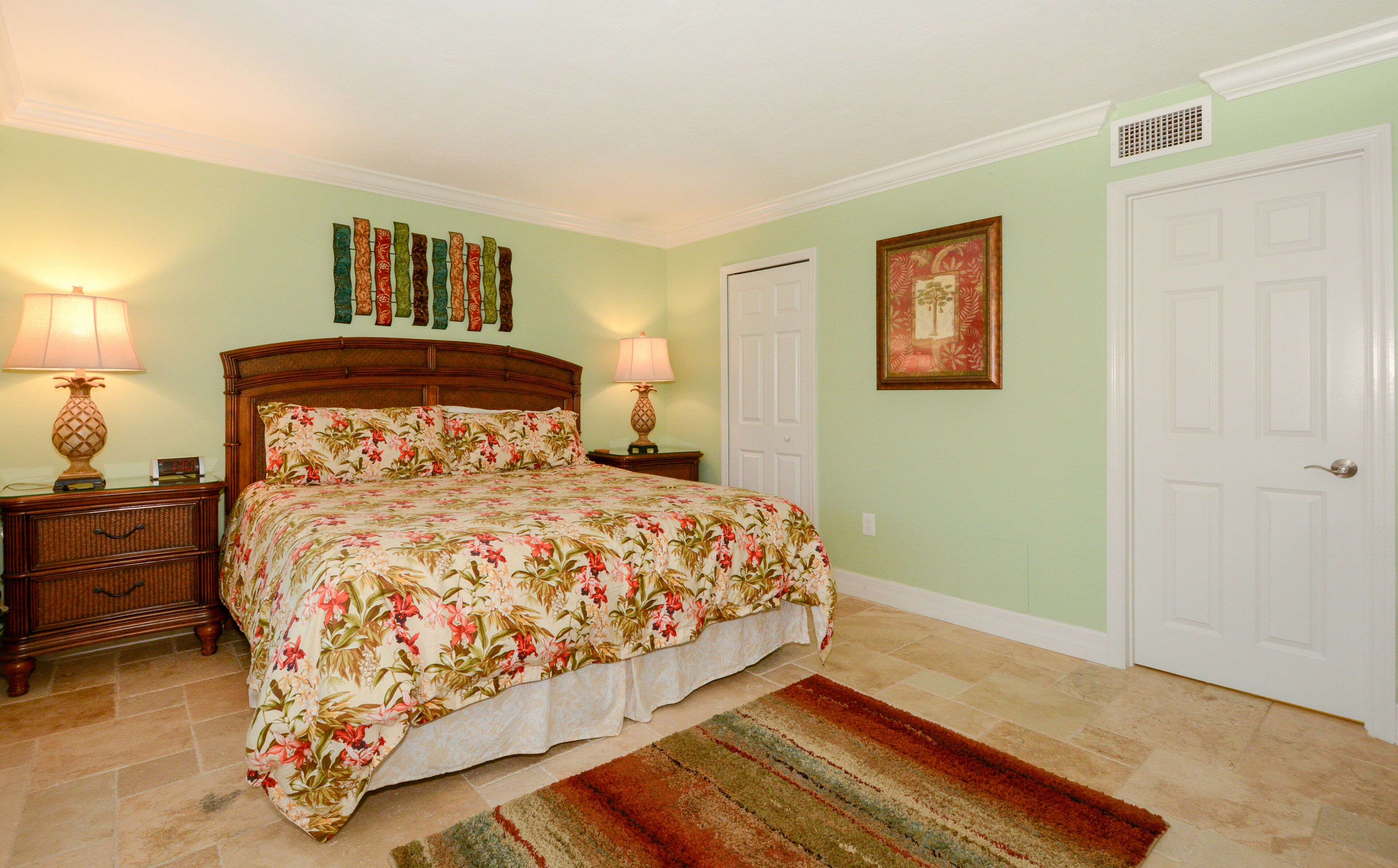 A bedroom with a floral bedspread, two bedside lamps, a nightstand, a rug, a door, and a wall with a framed picture.