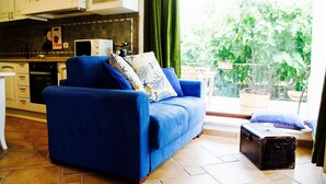 A living room with a blue sofa, a small table, and a suitcase. There is a kitchen area with cabinets and a window overlooking greenery. - TENUTA LE CRIE, VILLETTA RESIDENCE with swimming pool and tennis court (Muro Leccese)
