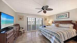A hotel room with a bed, a television, a ceiling fan, a window with blinds, and a view of the beach.
