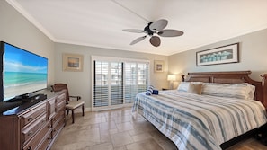 A hotel room with a bed, a television, a ceiling fan, a window with blinds, and a view of the beach.