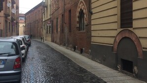 A cobblestone street lined with historic buildings, parked cars, and a pedestrian walkway. - Asti historic center cathedral (Asti)