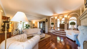 A spacious living room with a grand staircase, white leather sofas, and a chandelier. - Fontespina 6, Emma Villas (Conca degli Ulivi)
