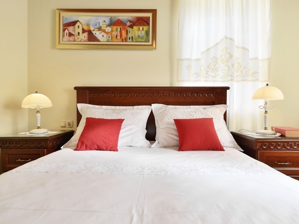 A neatly made bed with white linens and two red pillows, flanked by two bedside tables with lamps, in a room with a painting on the wall and a window with sheer curtains.