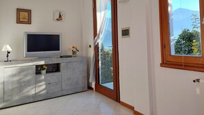 Un salon avec un téléviseur à écran plat posé sur un meuble gris, une petite table sur laquelle se trouve un vase de fleurs, et une porte donnant sur un balcon.