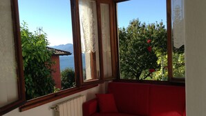 Une pièce avec un canapé rouge, une fenêtre ornée de rideaux en dentelle et une vue sur un arbre aux fleurs rouges, avec des montagnes en arrière-plan.