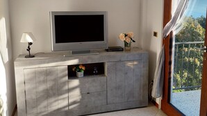 Un salon avec un téléviseur à écran plat posé sur un meuble gris, une petite table avec une lampe et un vase rempli de fleurs. Deux tableaux encadrés ornent le mur.