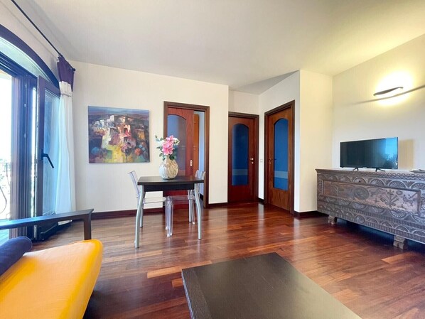 A modern living room with a wooden floor, a dining table with chairs, a sofa, a TV on a stand, and a painting on the wall. - Elegant apartment with sea view at La Maddalena (La Maddalena)