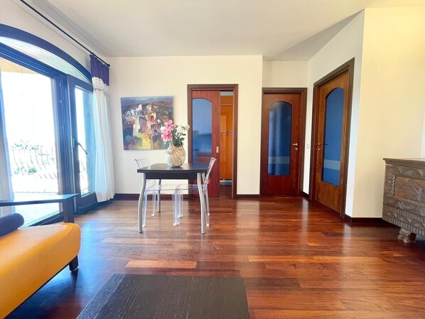 A spacious living room with a wooden floor, a dining table with chairs, a sofa, and a painting on the wall.
