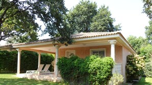 Eine überdachte Terrasse mit zwei Liegestühlen, ein Backsteinhaus mit Ziegeldach und üppigem Grün.