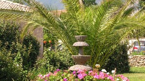 Ein Garten mit einem Steinfbrunnen, üppigem Grün und rosa Blüten.