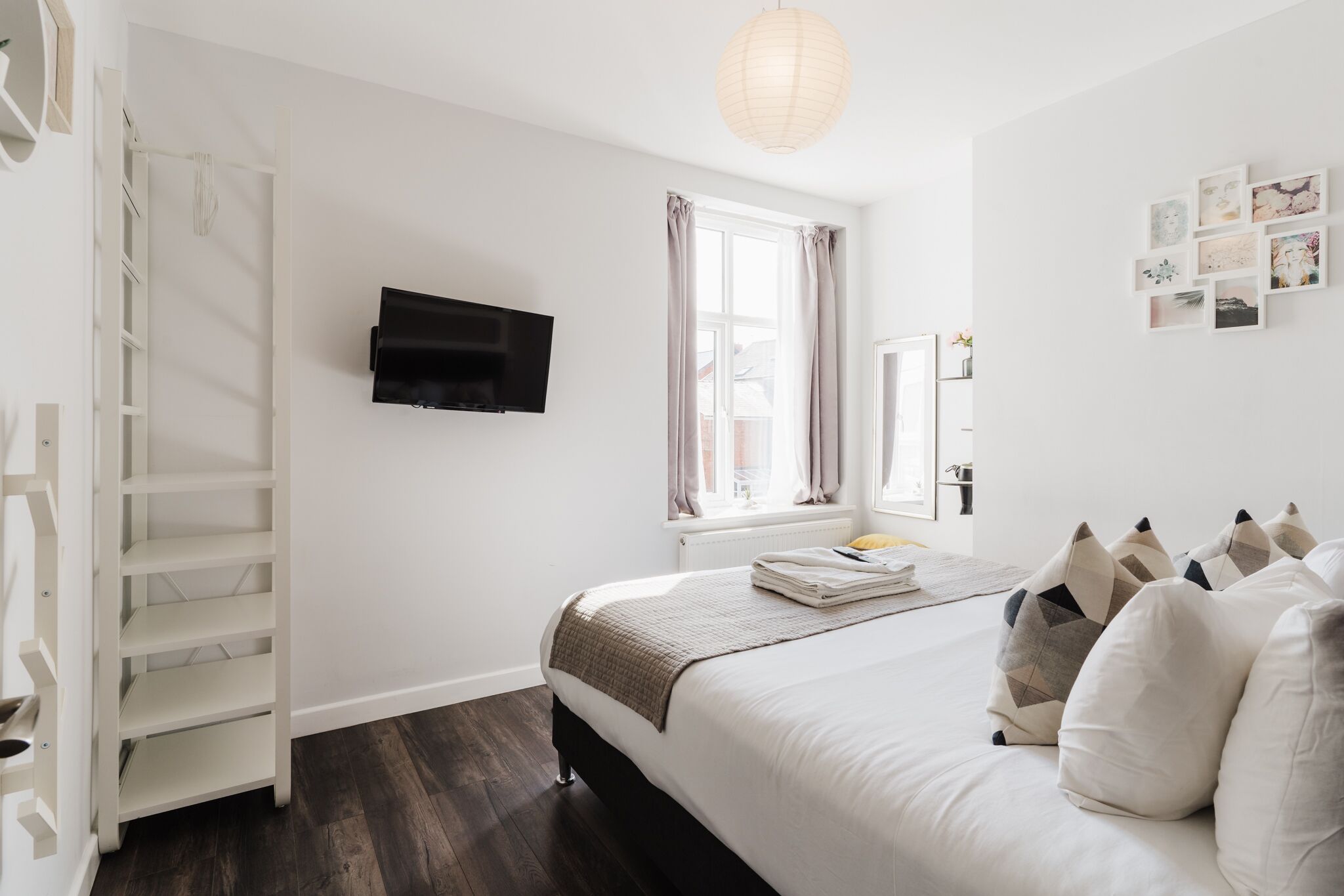 A bedroom with a bed, a TV mounted on the wall, a window with curtains, a shelf with pictures, and a white wardrobe.