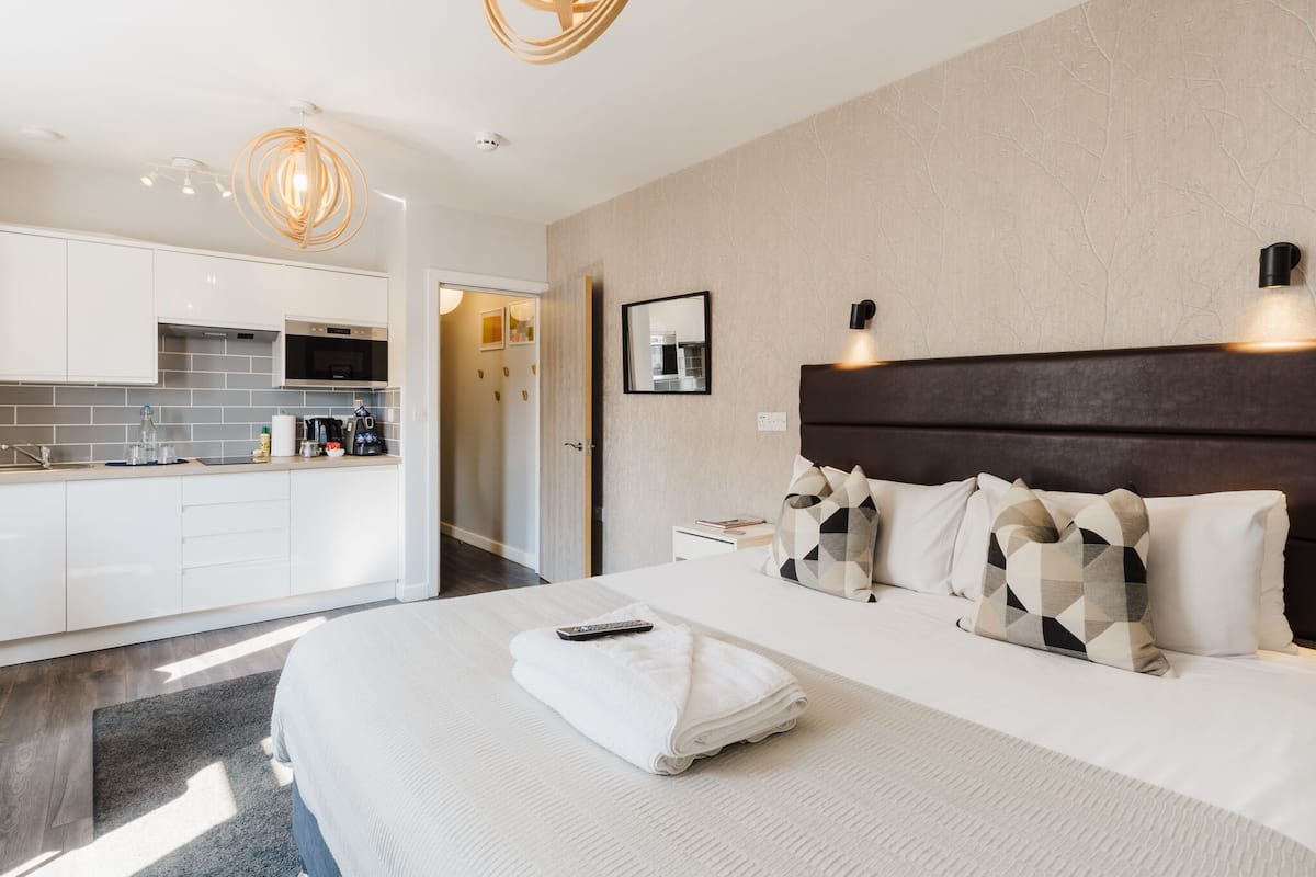 Ett modernt hotellrum med en stor säng, ett sängbord, en liten avlastningsyta, ett inbyggt kök med vita skåp och en lampa ovanför sängen.
