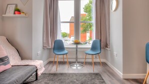 A modern room with a bed, two chairs, a small table, a window with curtains, a shelf with a picture and plants, and a clock on the wall. - Design Suites Lytham (Lytham St. Anne's)