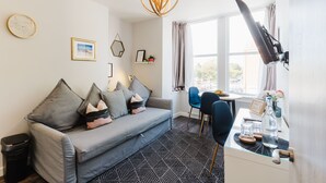 A modern living room with a grey sofa, a glass dining table, and a black chair. There is a window with curtains, a wall-mounted lamp, and a small shelf with decorative items. - Design Suites Lytham (Lytham St. Anne's)