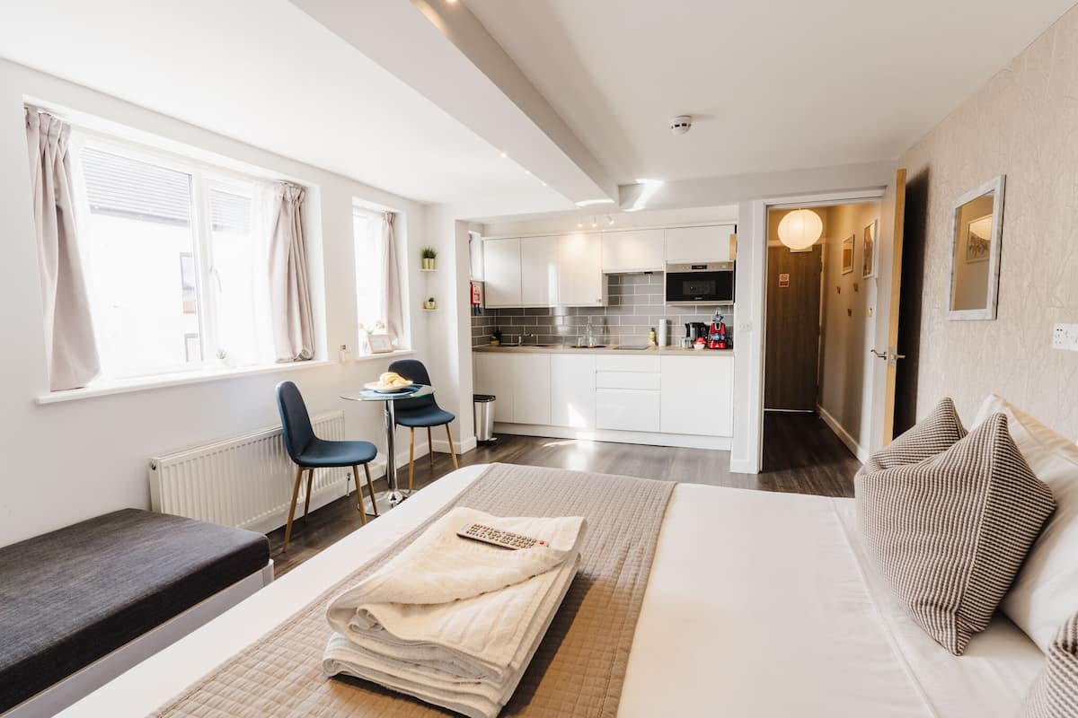 Ett modernt hotellrum med en säng, ett nattduksbord och utsikt över köket och vardagsrummet.