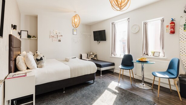 A modern hotel room with a bed, bedside table, two chairs, a small table, a TV, and a window with curtains. - Design Suites Lytham (Lytham St. Anne's)