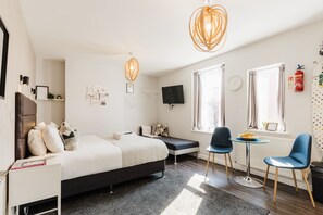 Quarto de hotel moderno com cama, mesa de cabeceira, duas cadeiras, uma pequena mesa, televisão e janela com cortinas.