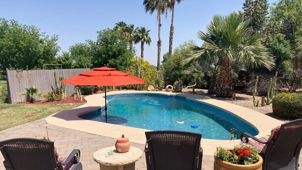 A swimming pool with a curved edge, surrounded by outdoor furniture including wicker chairs and a table, with a red umbrella and potted plants nearby. - Ranch Home with Guest Suite Located on 2 acres of Orchard (Waddell)