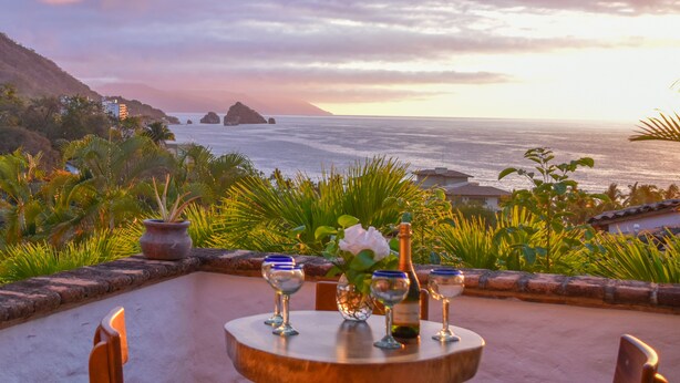 A table set with wine glasses and a bottle, overlooking a coastal view with palm trees and a mountainous backdrop. - Private villa- 3 bdrm & 4 bath with ocean views and new private infinity pool  (Puerto Vallarta)