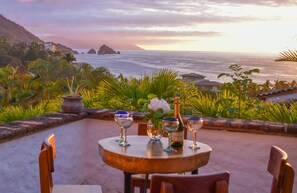 Mesa servida con copas de vino y una botella, con vista al mar, palmeras y montañas al fondo.