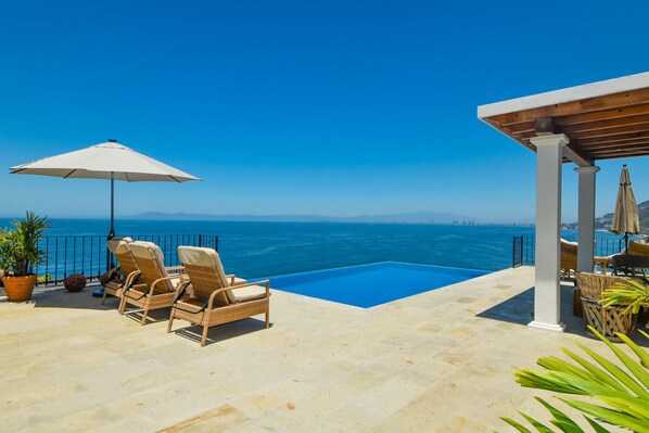 A poolside area with lounge chairs and an umbrella, overlooking a clear blue sea.
