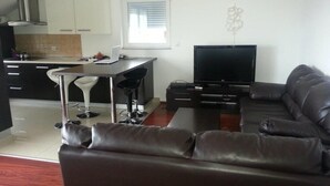 Ein modernes Wohnzimmer mit einem braunen Ledersofa, einem Flachbildfernseher, einem Essbereich mit einem weißen Stuhl und einer Küche mit weißen Schränken und einem Fenster.