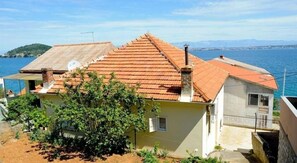 Situated in a coastal area, this property offers a view of the sea and distant hills, surrounded by a small garden with various plants.