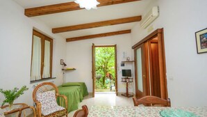 Ein Zimmer mit Holzbalkendecke, einem Esstisch mit floralem Tischläufer, Korbstühlen, einem grünen Sofa und einem Fernseher auf einem Schrank.