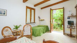 Ein Zimmer mit einem Bett, zwei Korbstühlen, einem kleinen Tisch mit einer Vase, einer grünen Tagesdecke, einem Holz-TV-Möbel und einem Fenster mit Blick auf Grünflächen.