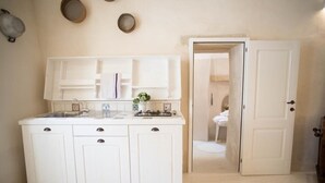 Eine kleine, weiße Küche mit Spüle, Herd und Schränken. Eine Tür führt zu einem Badezimmer, in dem eine Toilette zu sehen ist.