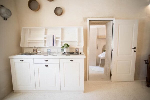 Une petite cuisine blanche, équipée d’un évier, d’une cuisinière et de placards. Une porte donne accès à une salle de bain où l’on peut apercevoir les toilettes.