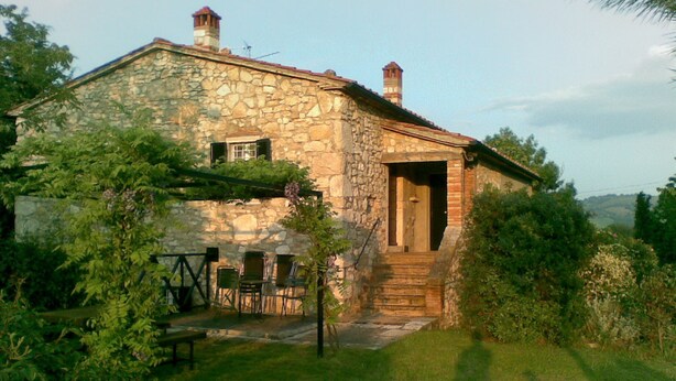 A stone house with a wooden door and steps leading to the entrance. There are chairs and a table outside.