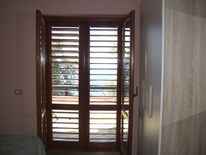 A room with a wooden door with horizontal blinds, a bed with a patterned bedspread, and a white wall with a light switch.
