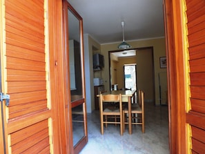 A room with wooden doors, a dining table with chairs, a ceiling fan, and a kitchen area with appliances.