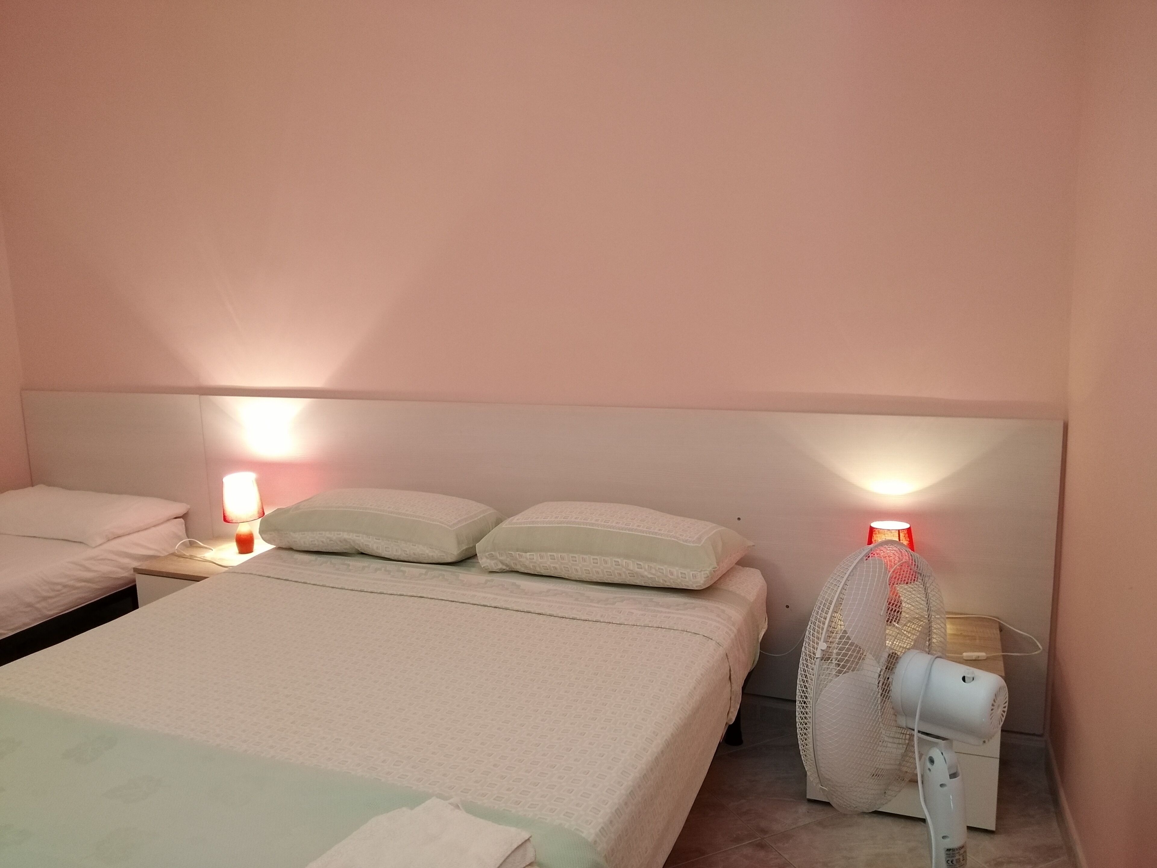 Camera d'albergo con due letti, ciascuno con una testiera e cuscini bianchi. Sono presenti due abat-jour con paralumi rossi, una ventola bianca e un riscaldatore bianco a pavimento.