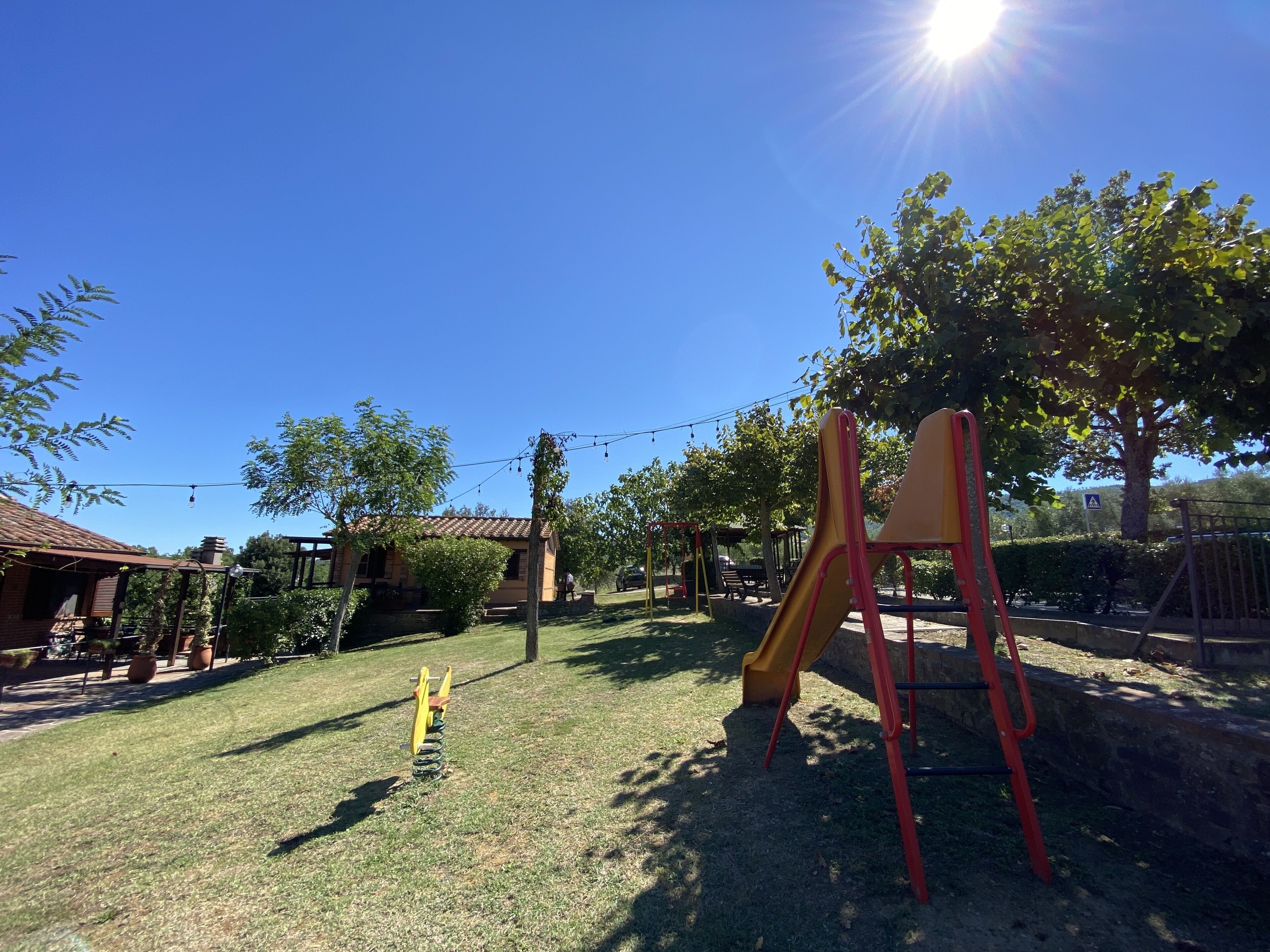 A playground with a slide and swings, surrounded by trees and a building with a tiled roof.