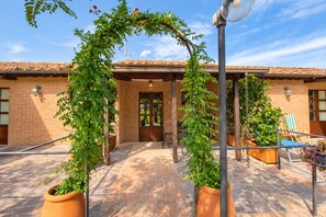 A courtyard with a brick building, potted plants, and outdoor seating. - Il Perugino | Agriturismo with covered patio (Paciano, PG)