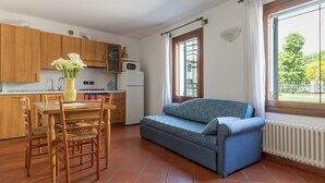 A compact kitchen with wooden cabinets, a dining table with chairs, a microwave, and a blue sofa near a window. - Apartment in farmhouse, Venice. Bedroom, bathroom, full kitchen / living room. (Oriago)