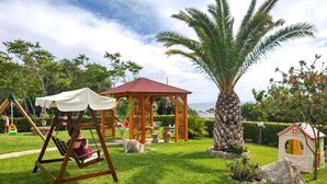 A garden with a swing set, a gazebo, a palm tree, and a small playhouse.