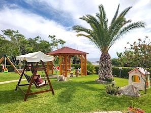 A garden with a swing set, a gazebo, a palm tree, and a small playhouse.