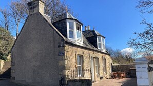 Une petite maison en pierre avec un toit à pignon très incliné, un petit porche et une enseigne indiquant « CARDY COTTAGE, RUE PRINCIPALE ».