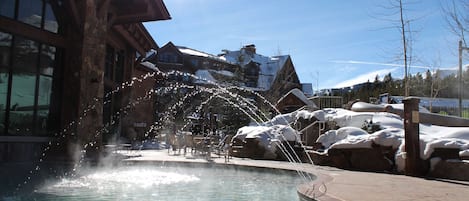 Una piscina con elementos acuáticos, un edificio de fachada de piedra y un cielo azul despejado.
