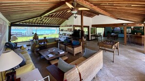 Un vaste espace de vie avec une grande fenêtre donnant sur une plage, des meubles en bois et une table de baby-foot.
