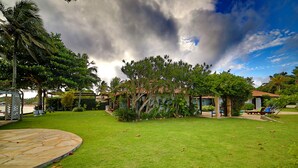 Un jardin tropical doté d’une terrasse en pierre, d’une végétation luxuriante et d’une maison avec un toit en tuiles.